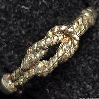 Close-up image of silver rope-style ring showing intertwined loop design and textured rope detail on black fabric background.