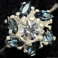 Close-up top view of the sterling silver ring showing the round clear center stone surrounded by six marquise-shaped blue stones and silver prong designs.