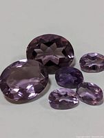 Six faceted genuine amethyst gemstones laid out on a white surface, showing different sizes and oval/round shapes with purple coloration.
