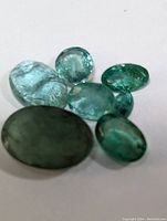 Six oval shaped genuine emerald gemstones in different green shades displayed on white background