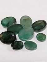 Nine loose genuine emerald gemstones of various shapes and shades of green laying on white background, showing size, shape, clarity, color variation, and surface details.