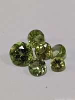 Six round faceted peridot gemstones arranged on a white background showing variations in size and green hue.