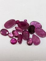 Close-up photo showing a collection of approximately 15 loose polished ruby gemstones in various shapes and sizes.