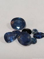 Seven loose sapphire gemstones of different shapes and sizes displayed on a white background showing various shades of blue.
