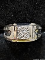 Close-up front view of silver ring with square face and diamond-style accents on black background.