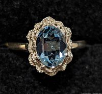 Close-up front of silver ring with blue oval faceted gemstone centered in ornate scalloped bezel setting against a black background.