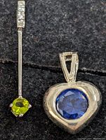 Close-up of both pendants showing the heart-shaped pendant with a large blue gemstone and the bar pendant with a small green gemstone and small clear stones on the bail.