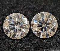 Two round brilliant cut clear gemstones placed on black fabric, showing faceted surfaces and sparkle.