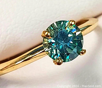 Close-up photo of the ring showing the round blue-green moissanite stone set in four prongs on a yellow gold band.