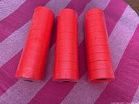 Photo of 15 rolls of bright orange plastic flagging tape stacked in two groups on a pink and purple striped fabric surface, tightly wound with visible creases.