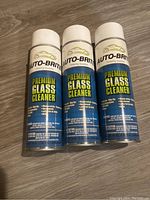 Three cans of Auto-Brite Premium Glass Cleaner arranged side by side on a wooden floor showing front of the cans.