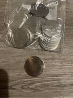 Plastic bag containing multiple silver metal circles, one metal circle removed and displayed in front showing the hole near edge and smooth surface.