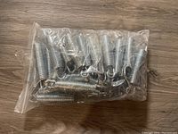 Bag containing multiple large metal coil springs with hooks on both ends lying on wooden floor.