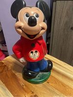 Front view of Mickey Mouse coin bank figure, showing the red shirt with Mickey Mouse Club logo and green base.