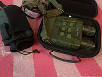 Night vision goggles inside the black protective carrying case with the olive green strap partly visible, resting on a pink and white striped fabric.