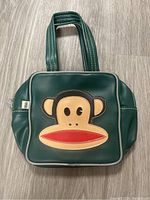 Front view of green Paul Frank handbag showing the Julius monkey face applique in tan, black and red with white piping and dual handles on a wooden floor.