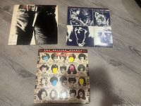 Three vinyl record album covers by The Rolling Stones laid out flat on a wooden floor. "Sticky Fingers" cover with iconic jeans zipper on the left, "Emotional Rescue" blue-toned art cover on the right, and "Some Girls" cover featuring illustrated faces with color highlights in the center bottom.