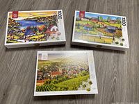 Three sealed 1000 piece puzzles showing different scenic artwork including coastal village, farm countryside, and vineyard town aerial view.