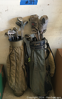 Photo of two golf bags with golf clubs inside against a wall. One bag is tan, the other green. Both have multiple clubs including irons and woods.