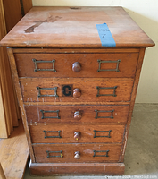 Front view of a five drawer wooden filing cabinet showing medium brown wood, round wooden knobs, metal label holders on each drawer, surface scratches and stains, and a blue tape tag on top.