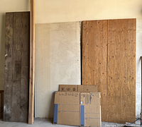 Photo showing one large plywood sheet, one large drywall piece, two 2x4 lumber pieces leaning vertically, and one decking board against the wall with flattened cardboard boxes in front (boxes excluded from sale).