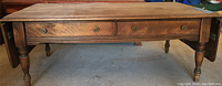 Full frontal view of wooden coffee table showing two drawers, drop leaves extended downward, and turned legs.