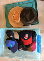 Top view of the plastic box containing two fedoras and multiple bill caps of various colors and logos.