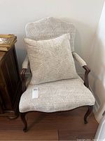 Full front view of the armchair with off-white upholstery, curved wood arms and legs, with matching throw pillow on seat.