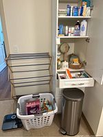 Photo of bathroom corner showing drying rack, laundry basket with cleaning supplies, a cabinet with toiletries, a bathroom mirror, and a stainless steel trash can