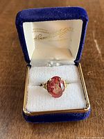 Ring shown in original blue velvet box, displaying the pink faceted synthetic stone and yellow 18K gold band with ornate details