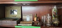 Shelf with old wooden box opened showing postcards and wooden clothespins, Taylor thermometers, glassware, keys, marbles in jar, small bottles, and razor case