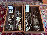 Overall view of two brown felt-lined wooden trays containing assorted costume jewelry pieces including pins, necklaces with pendants, bracelets, and earrings.