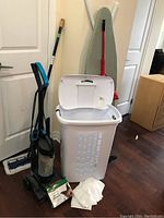 Full lot view showing Bissell vacuum, laundry basket with lid open, broom, mop, ironing board and garbage bags