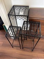 Set of three decorative black metal plant stands shown together from above and front angle highlighting scrollwork tops and legs.