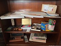 Photo showing shelves with several paper pads, wooden crafting tools, a metal case with colored markers or pens, collection of ink stamps, a can with scissors, and other miscellaneous craft supplies.