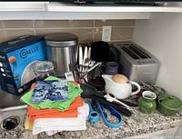 Overview of the lot showing Corelle Classics box, toaster, waste can, various cutlery, ceramic teapot, green cream and sugar bowl, mugs, kitchen tools, and fabric towels on granite countertop.
