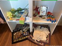 View of shelving unit with adult coloring books, floral artificial stems, boxed drawer liners on upper left; small hooked rug with house and tree scene, folded fine linens and smaller rug on lower left; assorted handcrafted items including origami crane garland, framed print, beaded flying goose and crocheted doily on right compartments.