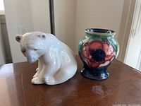 Side view of seated glossy white polar bear figurine and small floral Moorcroft jar on wooden table near window.