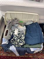 Wide view of folded Olsen ladies clothing items including striped tops, floral blouse, knit sweaters, and denim look pants stacked in a plastic bin.