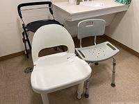 Photo showing two shower chairs (white and gray) and a red and black TRUST walker with a tray in a bathroom