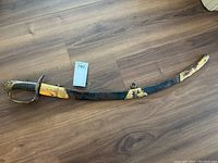 Full-length view of curved blade in brass and leather scabbard, on wood floor