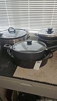 Three cooking pots and pans with glass lids arranged together including a rectangular and round pan, all in good condition.