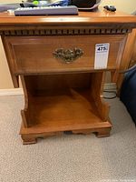 Front view of wooden bedside table showing single drawer with ornate metal handle and open shelf underneath.