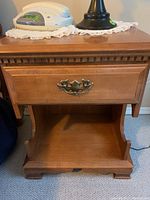 Front view of wooden bedside table showing single drawer with ornate metal handle and open bottom shelf.