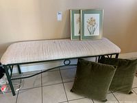 Full view of metal framed bench with beige upholstered seat, two framed botanical prints resting on seat and two green cushions on floor in front. Shows overall condition and style.