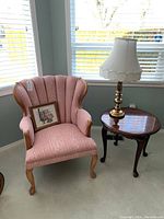 View of pink upholstered armchair with scalloped back, wooden frame and legs, wooden oval side table to the right, and brass table lamp with white fabric shade on the table.