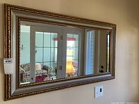 Full front view of the decorative mirror hanging on a beige wall reflecting a room with a sofa, lamp, and a glass door.