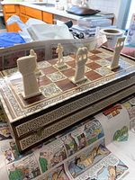 Chess set showing ivory-colored chess pieces placed on the wooden and mother of pearl inlaid board on a table covered partially with newspaper comics.