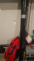 Wide view of fishing rods standing vertically next to red and blue life jacket vest and a gray tackle box.