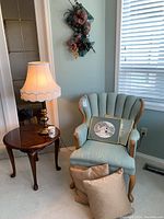 View of the soft green upholstered armchair next to the dark wood side table with brass lamp on top, two brown pillows on floor, and framed artwork on chair.